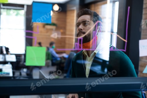 Preview: Mixed race man wearing face mask looking at information on glass wall in office