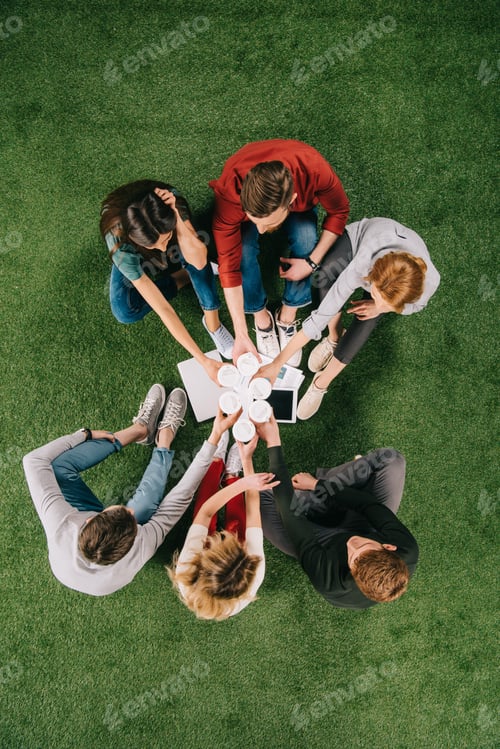 Preview: overhead view of business colleagues clinking cups with coffee while sitting on grass