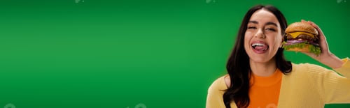 Preview: joyful woman with delicious burger sticking out tongue and looking at camera isolated on green,