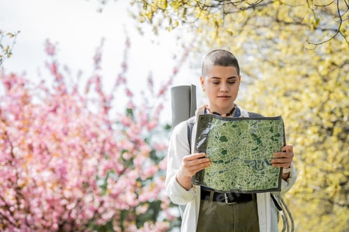 Preview: Young short haired female tourist in casual clothes with backpack looking at map while standing with