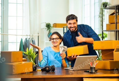Preview: Happy Indian entrepreneurs show cash and packed boxes in warehouse after successful sales