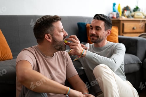 Preview: Gay couple sharing grapes and wine at home, celebrating love and lgbtq+ pride