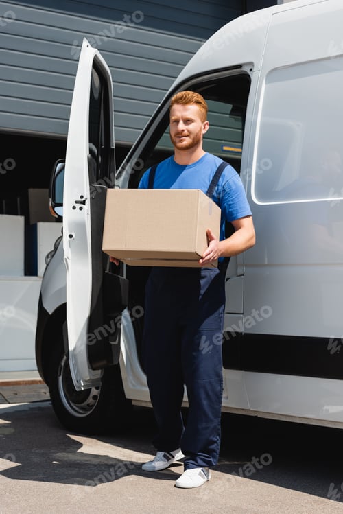 Preview: Young loader looking away while holding carton box near truck on urban street