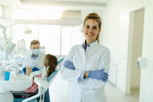 Preview: Portrait of young smiling female dentist posing with crossed arms while standing in dental office.