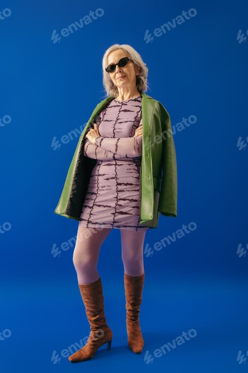 Preview: elegant grey haired woman in suede boots and green leather jacket over purple dress standing with
