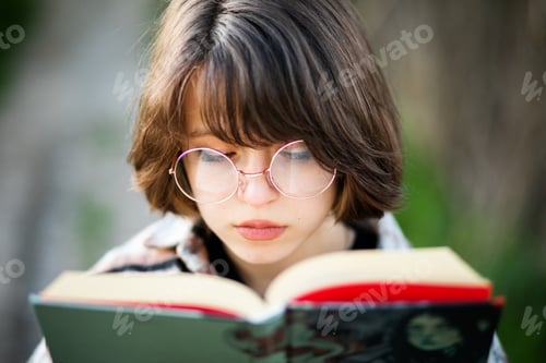 Preview: a teenage girl with short dark hair in glasses holds a book in her hands close to her face.