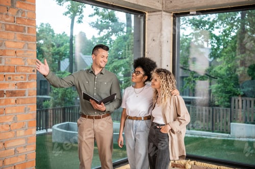 Preview: smiling realtor pointing with hand and showing interior of new home to interracial lesbian couple