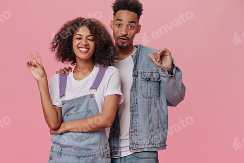 Preview: Cheerful girl showing peace sign while her boyfriend smiling on pink background. Pretty woman in wh