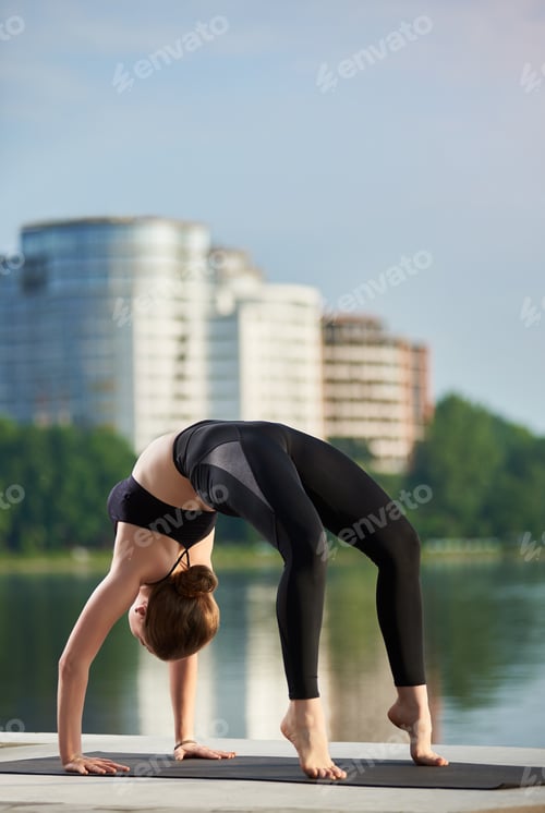 Preview: Slender girl practicing yoga outdoors near the river, standing in Upward bow Wheel pose