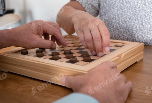 Preview: Two senior people at home while playing a game of checkers on wooden table.