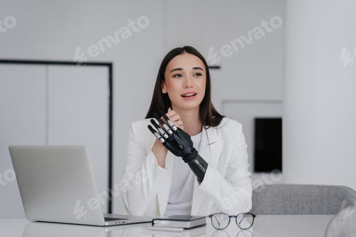 Preview: brunette caucasian woman in white suit with artificial arm prosthesis sitting at desk with laptop
