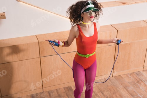 Preview: stylish woman in cap and body suit exercising with skipping rope