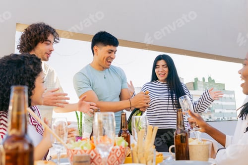 Visualização: Jovem latino abrindo champanhe em uma reunião ao ar livre em um terraço com amigos, animado
