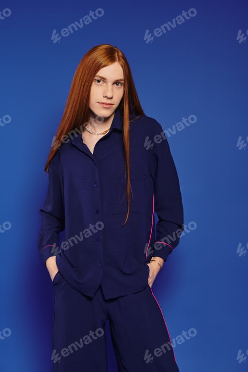 Preview: Stylish young man with long red hair poses confidently against a vibrant blue backdrop