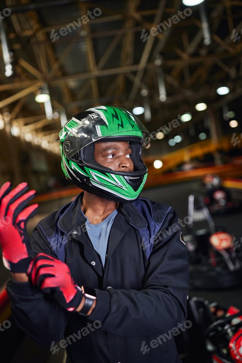 Preview: african american motorsports driver in helmet wearing gloves and standing in karting track