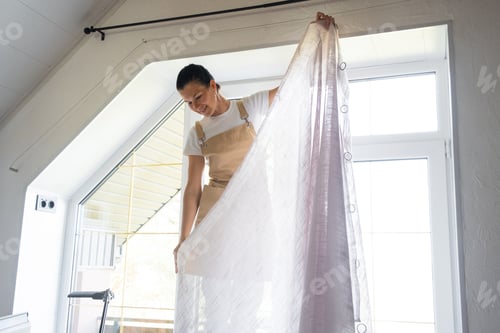 Preview: Woman in apron hangs transparent tulle curtains on large attic windows