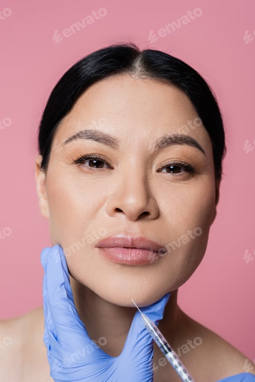 Preview: Cosmetologist holding syringe near chin of asian woman looking at camera isolated on pink
