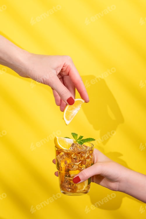 Preview: A hand holds a glass of iced tea with lemon and mint on a yellow background with shadow.