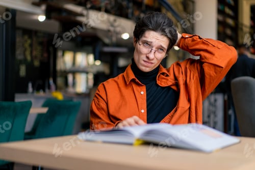 Preview: tired boy student sitting in front of a book in the library bored and lazy while preparing for exams