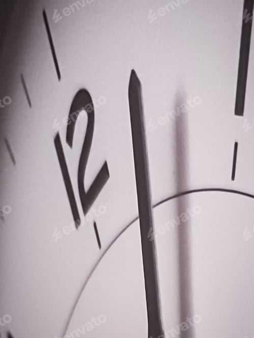 Preview: Closeup of the minute hand on a white clock face passing the number twelve