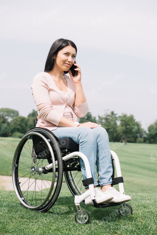 Preview: happy disabled woman in wheelchair talking on smartphone while resting in park
