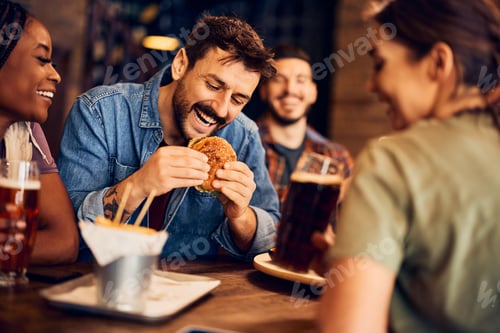 Preview: Happy man eating burger while gathering with friends in a bar.