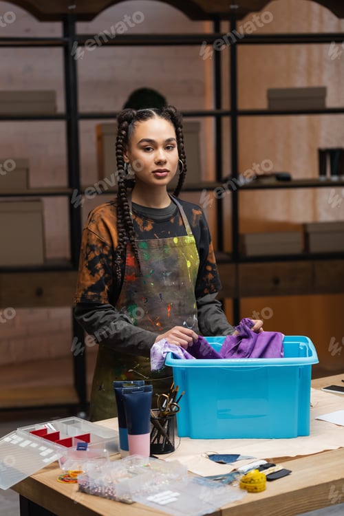 Preview: African american designer holding cloth near box and looking at camera in workshop