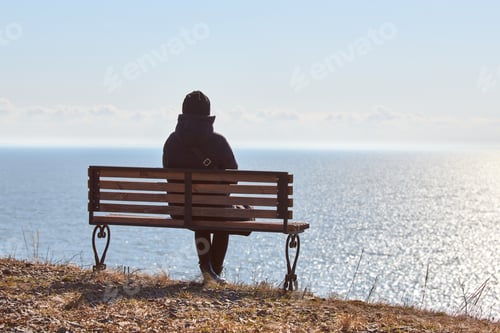 Preview: Single girl in a black jacket and hat sitting on bench at cliff at front of sea peaceful quiet place