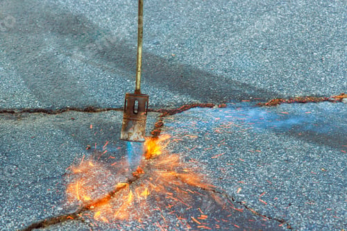 Preview: An asphalt patcher performs restoration work in cracks of road surface by burning dry grass before