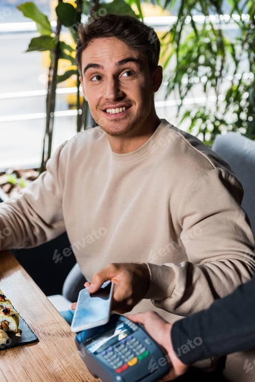 Preview: cropped view of waitress holding payment terminal near happy man with smartphone