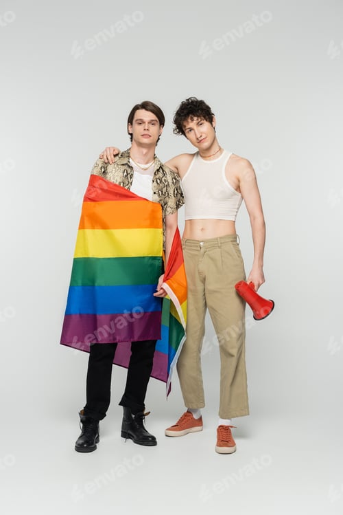 Preview: full length of young nonbinary couple with lgbt flag and megaphone looking at camera on grey