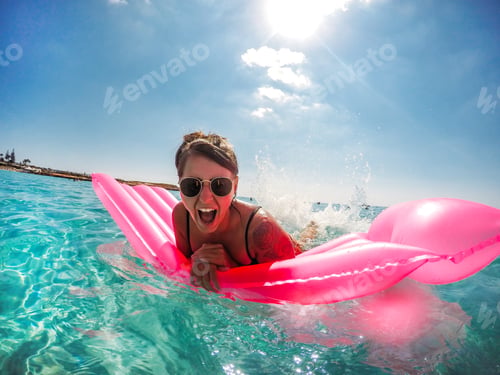 Preview: Happy smiling laughing young woman splashing in ocean on a pink inflatable in the summer