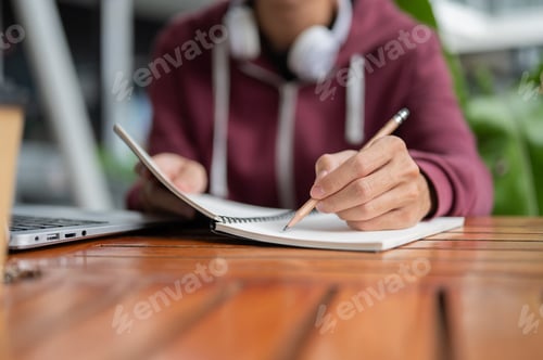 Preview: Close up college student man writing in notebook with a pencil aside laptop sitting at table in cafe