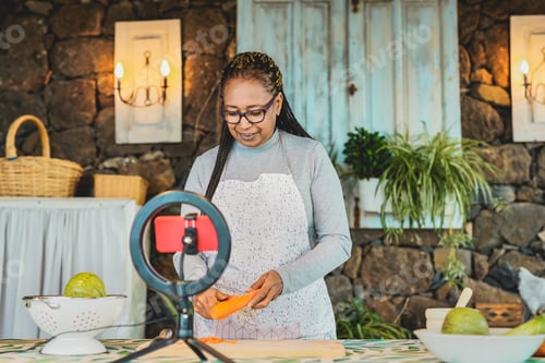 Preview: Cheerful african senior woman teach how to cook while streaming live video with smartphone outdoor