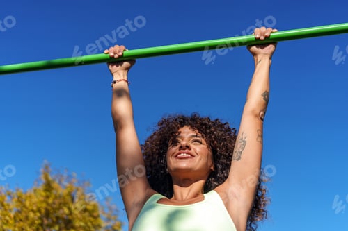 Preview: Happy young woman training arms on outdoor pull up bar, enjoying street workout under a blue sky