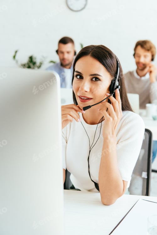 Preview: selective focus of attractive broker touching headset near coworkers in office
