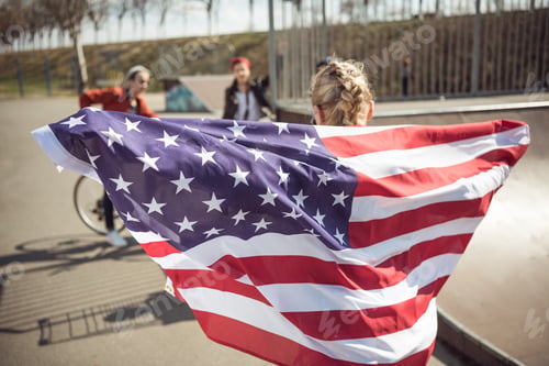 Preview: Back view of young girl standing with american flag and looking at friends at skateboard park
