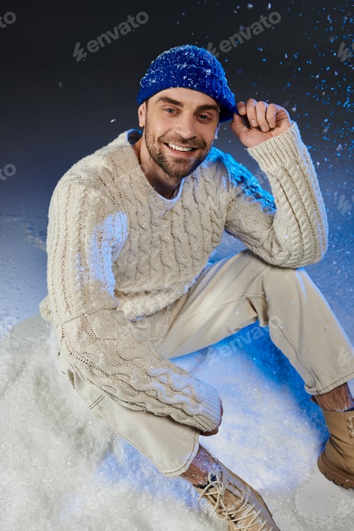 Preview: Joyful young man in a cozy sweater and blue beanie enjoying winter in a snowy setting
