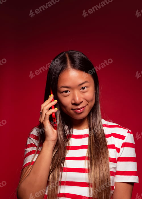 Preview: young chinese woman smiling and talking on smartphone while standing on red background
