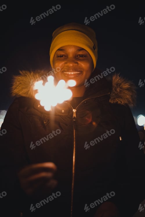 Preview: A young woman holding a sparkler at night with lights in the background