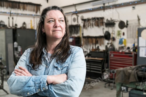 Preview: Woman with brown hair wearing Denim shirt standing in metal workshop, arms folded, smiling.
