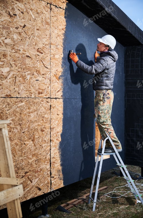Preview: Male painter using paint roller, doing exterior paint work while building wooden frame house.