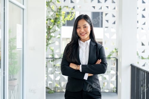 Preview: A confident Asian businesswoman stands in a corridor with her arms crossed, smiling at the camera.