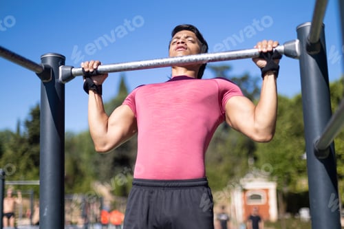 Preview: Man in pink shirt doing pull-ups on outdoor gym equipment in a park, focusing intensely.
