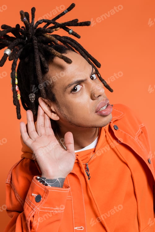 Preview: tattooed and multiracial man with dreadlocks looking at camera while touching ear isolated on coral