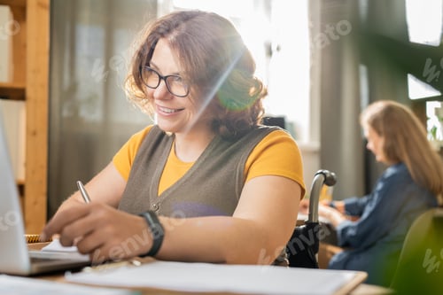 Preview: Young smiling disable office manager in casualwear looking at laptop display