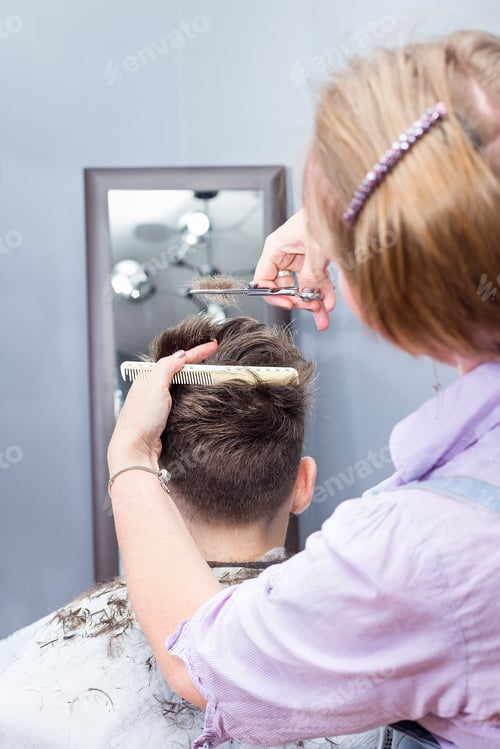 Preview: Nine years boy getting haircut by barber or hairdresser, sitting in chair at barbershop, vertical