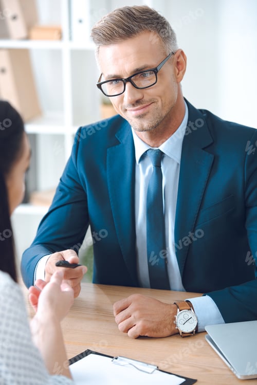 Preview: smiling insurance agent giving pen to female client at tabletop in office