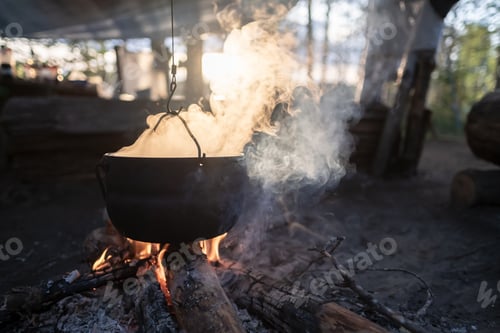 Preview: Bowler with escaping steam hangs over campfire, food is prepared at sunset, against of lake