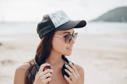 Preview: attractive woman in cap and sunglasses with headphones at beach